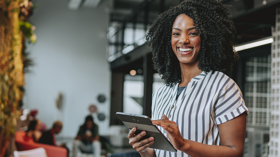 Foto de uma mulher negra em um escritório. Ela está segurando um tablet e demonstrando felicidade por saber do novo Selo GPTW para empresas que valorizam os jovens aprendizes.