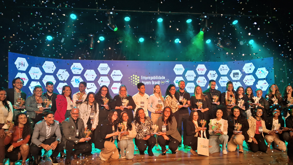 Foto de grupo registrada no palco do Teatro CIEE, em São Paulo, durante a entrega do Troféu Empregabilidade Jovem Brasil 2025. Aproximadamente 40 pessoas estão alinhadas em duas fileiras, sorrindo e segurando seus troféus, que têm o formato de peças de quebra-cabeça coloridas. No fundo, um painel azul exibe os logotipos das empresas homenageadas. Confetes prateados estão caindo, registrando o clima de celebração. A iluminação do palco é azul esverdeada, dando destaque ao cenário.