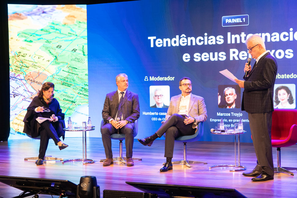 Quatro pessoas em cima de um palco, uma mulher à esquerda e três homens ao centro/ direita. A iluminação do palco é roxa e azul, todos usam vestes sociais.