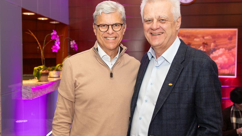 Dois homens estão em pé e sorrindo para a câmera. À esquerda está Robert John van Dijk, de cabelos grisalhos e óculos, vestindo suéter bege. À direita está Humberto Casagrande, de cabelos brancos, vestindo blazer azul escuro e camisa social azul clara. Ao fundo, parede de madeira com uma pintura e um relógio redondo.