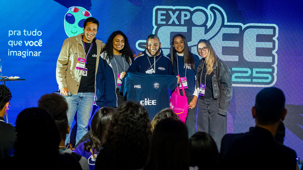 Aproximadamente cinco pessoas em um palco, a do meio segura uma blusa do CIEE