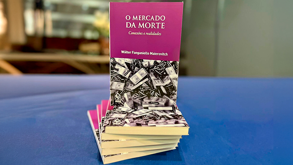 Imagem de um exemplar do livro “O Mercado da Morte: Conexões e Realidades”, de Wálter Fanganiello Maierovitch, empilhado sobre outros exemplares em uma mesa azul. A capa é roxa na parte superior e exibe uma imagem de notas de dinheiro e munições na parte inferior.