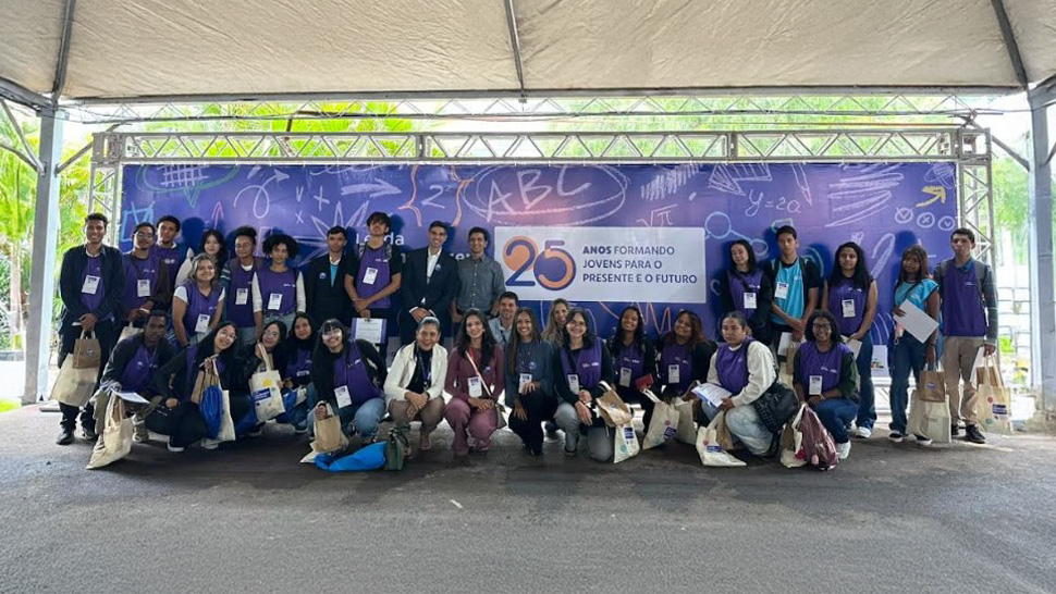 Um grupo de jovens reunidos em frente a um painel roxo com elementos gráficos de educação e o selo de 25 anos da Lei da Aprendizagem. Há uma placa ao centro, onde está escrito: "25 anos formando jovens para o presente e o futuro."