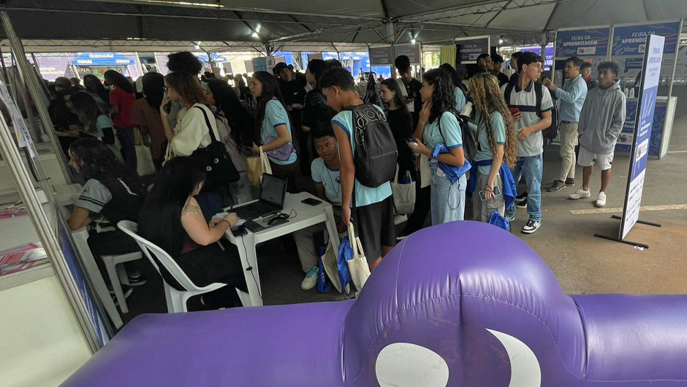Diversos adolescentes e jovens formam fila e recebem atendimento no estande do CIEE. À frente, um inflável roxo com o logo do CIEE ocupa a parte inferior da imagem. Ao fundo, profissionais orientam os participantes sobre cadastro, vagas de Estágio e Aprendizagem.