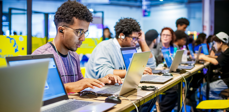 Grupo de jovens concentrados em frente a notebooks, participando de uma atividade formativa em tecnologia. A cena acontece em um ambiente colorido e colaborativo, com fones de ouvido e materiais de estudo.