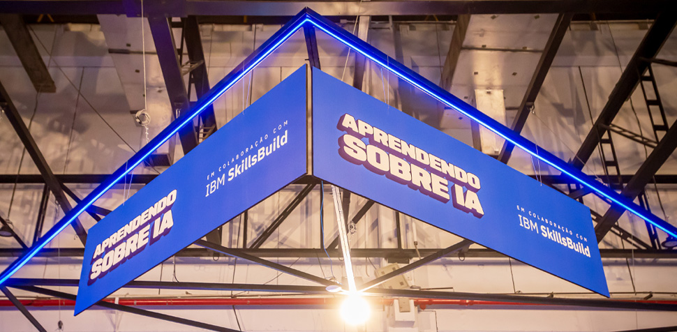 Estrutura triangular azul pendurada no teto de um evento, com a frase “Aprendendo sobre IA” em destaque e o logotipo do IBM SkillsBuild. O ambiente é iluminado e apresenta uma estética tecnológica e moderna.