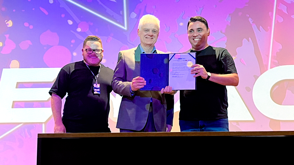Na imagem, três homens estão em pé sobre um palco iluminado com luzes roxas e rosadas. O homem ao centro, de cabelos brancos e terno cinza, segura um certificado aberto ao lado do homem à direita, que veste camiseta preta e jeans. À esquerda, outro homem também de camiseta preta sorri para a câmera.