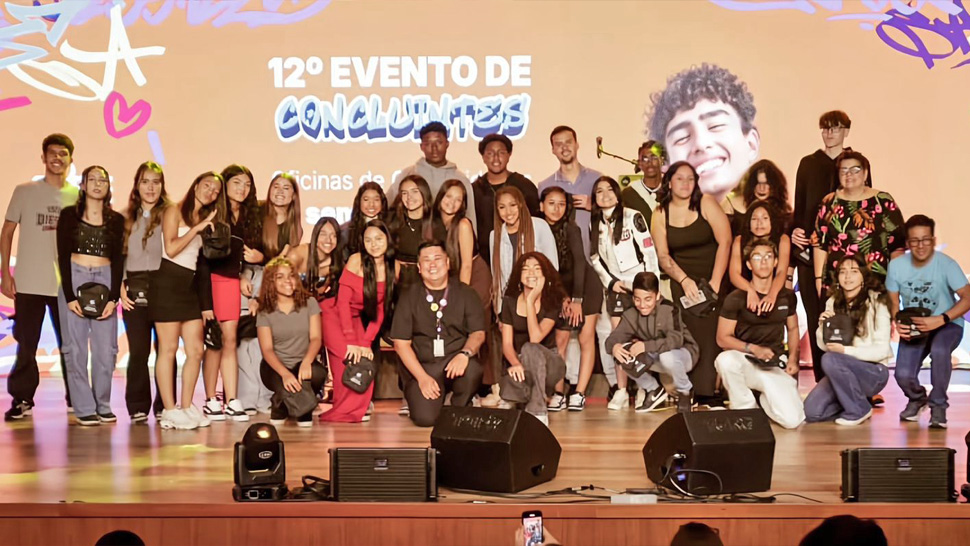 Foto de um grande grupo de jovens participantes das Oficinas de Cri@tividades posando no palco do Teatro CIEE durante o 12º Evento de Concluintes. Eles estão sorrindo, alguns fazendo poses descontraídas, enquanto ao fundo aparece o telão colorido com artes gráficas e o nome do evento. O clima é de celebração, alegria e conquista coletiva.