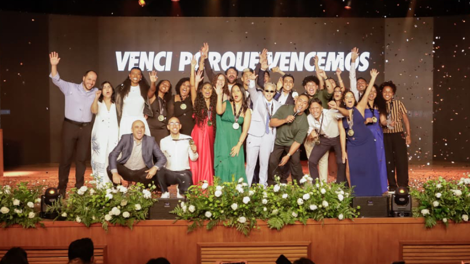 Grupo de formandos do projeto Somos CIEE posa no palco durante a cerimônia de formatura. Eles estão sorrindo, alguns com as mãos erguidas em comemoração. Ao fundo, um telão exibe a frase “Venci porque vencemos”.
