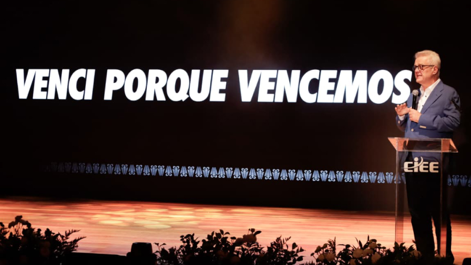 Humberto Casagrande, CEO do CIEE, aparece no palco durante a cerimônia, posicionado ao lado de um púlpito transparente com o logo do CIEE. Ele usa terno azul e fala ao público enquanto, ao fundo, um telão exibe a frase “Venci porque vencemos”.