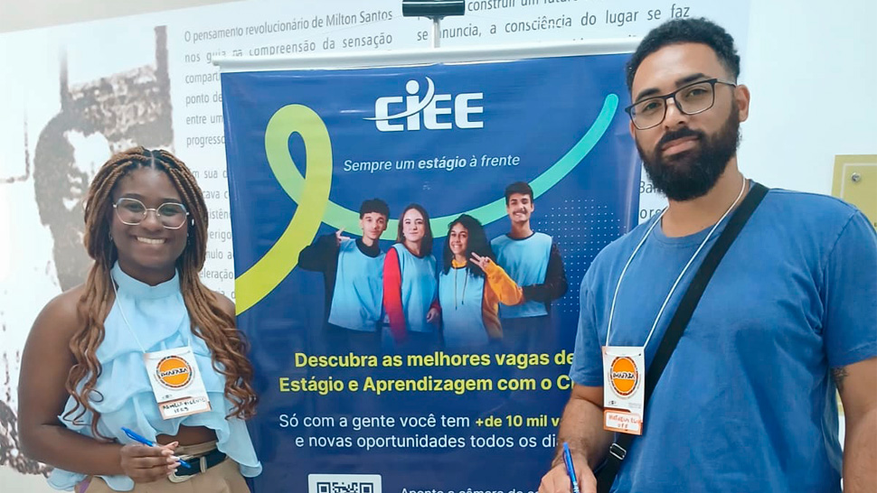 Dois jovens participantes posam em frente a um banner do CIEE. À esquerda, uma jovem negra de cabelos trançados, óculos e blusa azul clara sorri segurando uma caneta. À direita, um jovem negro de barba, óculos e camiseta azul escura também segura uma caneta.