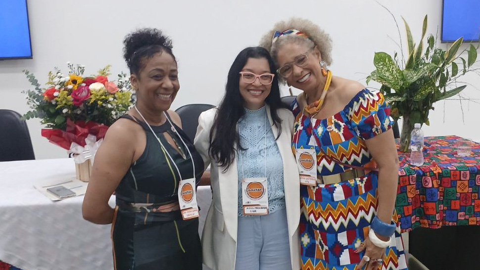 Três mulheres sorrindo posam lado a lado em um ambiente de evento. A mulher à esquerda usa um vestido preto com detalhes em verde; ao centro, uma mulher com blazer branco e blusa azul clara; e à direita está Sônia Guimarães, usando um vestido colorido estampado em tons vibrantes, acessórios grandes e faixa no cabelo.