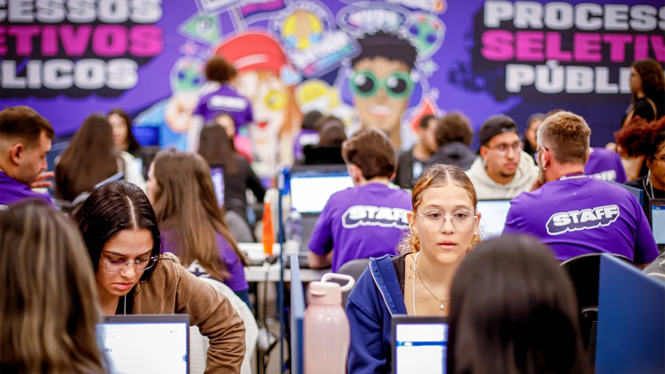 Jovens participam de entrevistas para encaminhamento de vagas oferecidas pelo CIEE, sentados frente a frente em mesas com computadores. Ao fundo, pessoas com camisetas roxas escritas "STAFF" auxiliam os participantes. A parede é decorada com ilustrações coloridas e a frase “Processos Seletivos Públicos”.