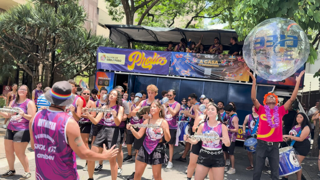 Foto de um bloco de Carnaval de rua durante o dia, com várias pessoas tocando instrumentos de percussão e chocalhos enquanto caminham. Ao fundo há um trio elétrico com faixas do bloco Pholia e menção ao CIEE.