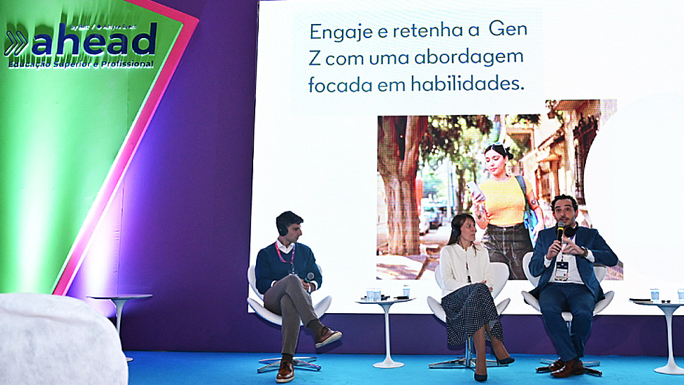 Três palestrantes sentados em poltronas brancas em um palco com iluminação verde e roxa durante o Fórum Ahead. Ao fundo, um telão exibe a frase “Engaje e retenha a Gen Z com uma abordagem focada em habilidades”, acompanhada da imagem de uma jovem caminhando na rua e um QR Code ao lado direito. Um dos palestrantes segura um microfone enquanto fala.