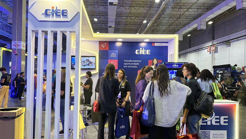 Estande do CIEE com estrutura iluminada nas cores da marca e logotipo em destaque. No espaço, várias pessoas interagem, conversam e recebem atendimento, algumas segurando brindes e sacolas. Ao fundo, painéis institucionais e telas reforçam a comunicação visual, enquanto o ambiente amplo do pavilhão mostra a movimentação intensa do evento.