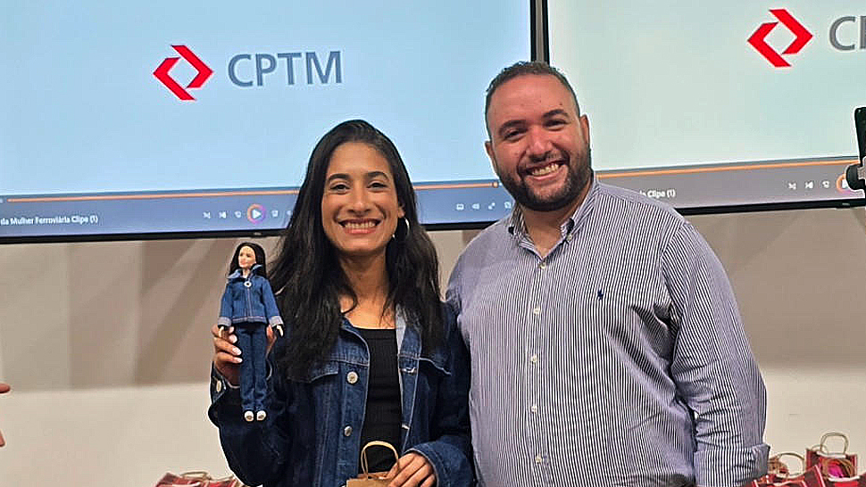 Bruno Melo, coordenador de Operações e Atendimento da Administração Pública do CIEE, ao lado de uma funcionária do quadro da CPTM, em frente a um telão com o logotipo da empresa. A colaboradora segura uma boneca Barbie personalizada com uniforme ferroviário, representando profissionais homenageadas no evento.