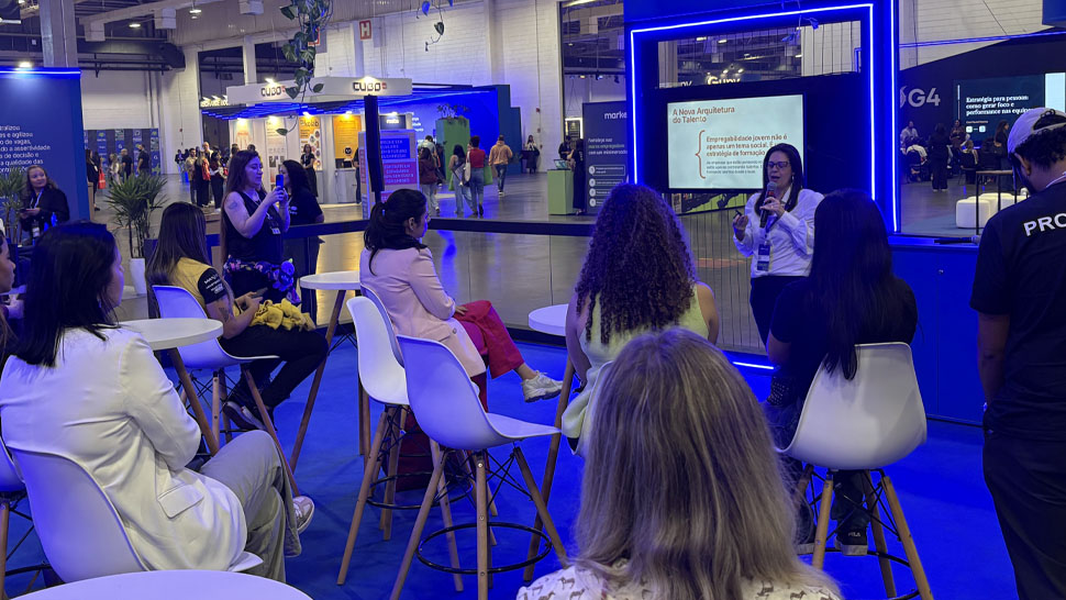 Palestra realizada durante evento, com Mônica Vargas, superintendente Nacional de Operações e Atendimento em pé, apresentando conteúdo ao público. À sua frente, participantes estão sentados em cadeiras altas, acompanhando a apresentação. Ao fundo, uma tela exibe o tema relacionado ao futuro do trabalho, enquanto o ambiente do pavilhão mostra outros estandes e circulação de pessoas.