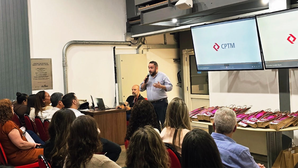 Bruno Melo, coordenador de Operações e Atendimento da Administração Pública do CIEE, fala ao microfone durante evento da CPTM para uma plateia sentada em auditório. Ao fundo, um telão exibe o logotipo da CPTM e, ao lado, há uma mesa com diversas sacolas de presentes organizadas. Participantes acompanham a apresentação sentados em cadeiras vermelhas.