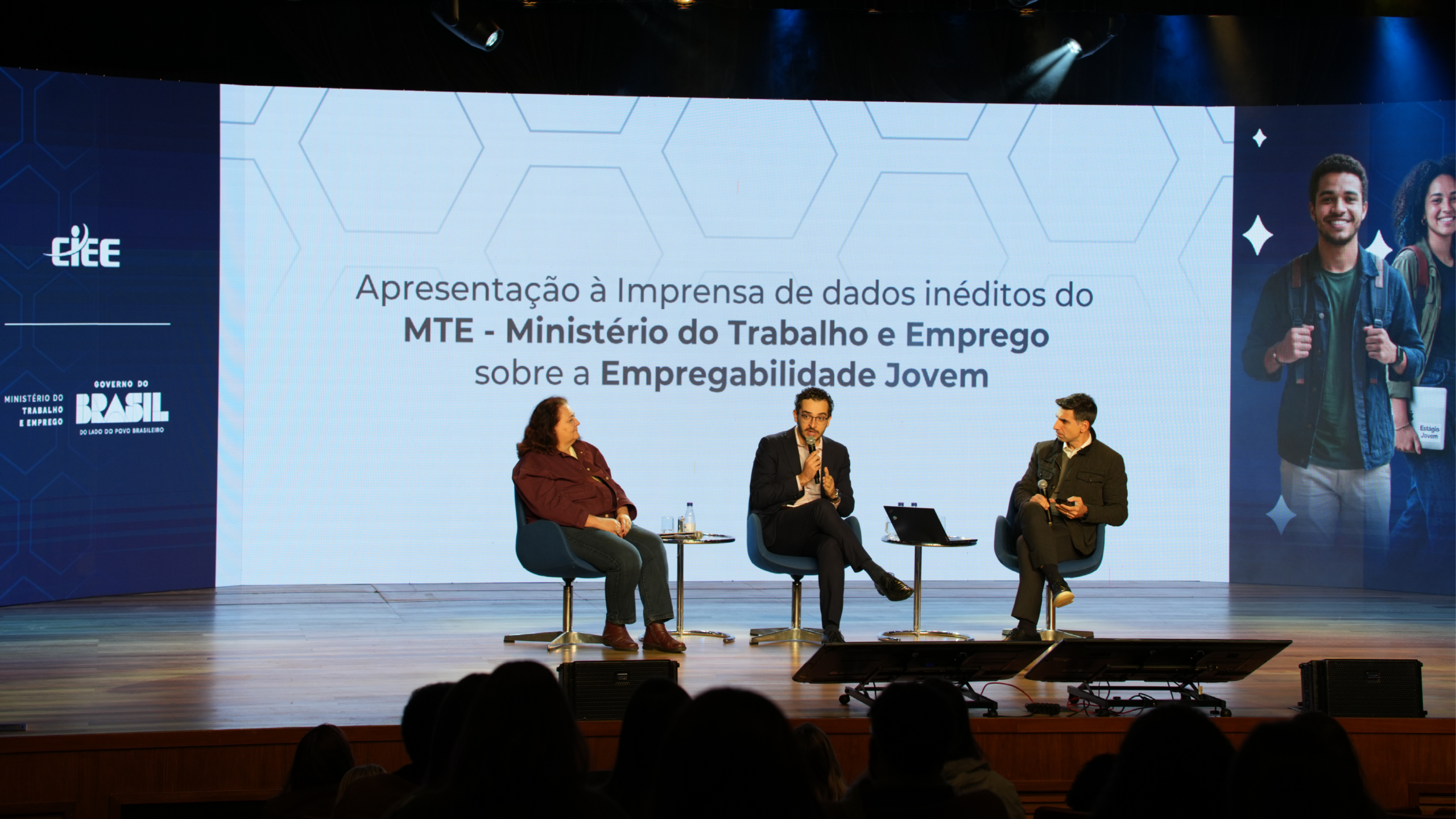 Fotografia horizontal de um evento em um palco de madeira bem iluminado. Três pessoas estão sentadas em poltronas azuis participando de um painel. Ao centro, um homem de terno escuro e óculos fala ao microfone. À esquerda dele, uma mulher de camisa bordô e calça jeans o escuta atentamente. À direita, um homem de jaqueta escura segura um microfone e um celular, também prestando atenção. Entre as poltronas, há pequenas mesas redondas de vidro apoiando copos d'água e um notebook. Atrás deles, um grande telão ocupa o fundo do palco. No centro, sobre um fundo claro com padrão de hexágonos, está escrito: "Apresentação à Imprensa de dados inéditos do MTE - Ministério do Trabalho e Emprego sobre a Empregabilidade Jovem". Nas laterais azuis escuras do telão, estão os logotipos do CIEE e do Ministério do Trabalho e Emprego (Governo do Brasil) à esquerda, e uma foto de um rapaz e uma moça jovens e sorridentes, com mochilas e cadernos, à direita. Em primeiro plano, na parte inferior da foto, veem-se as silhuetas escuras das cabeças das pessoas que formam a plateia.