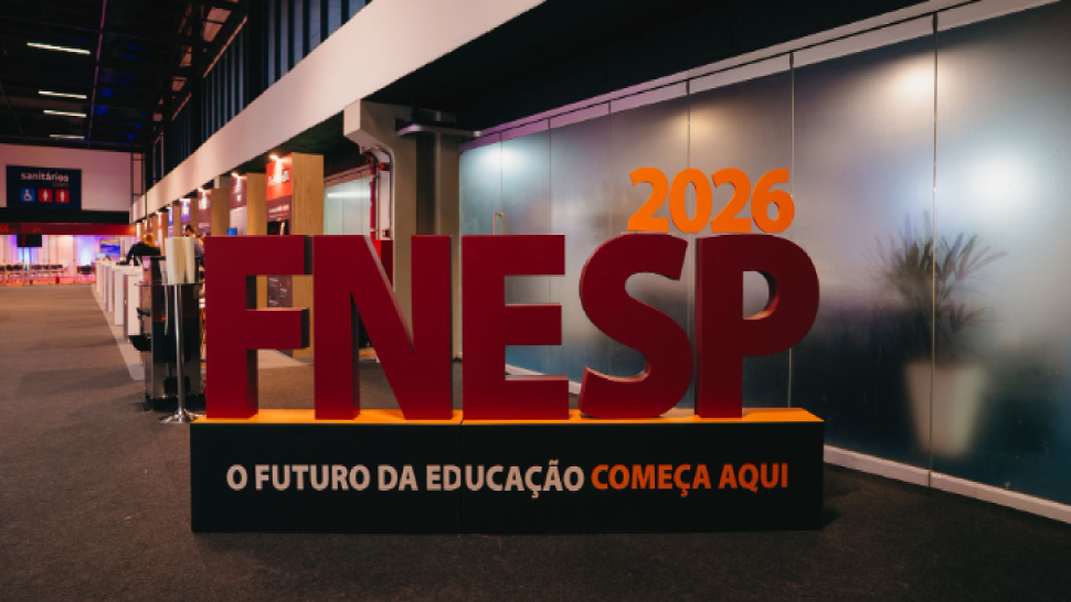 Fotografia de um grande letreiro 3D em um pavilhão de eventos. Em destaque no centro, a sigla "FNESP" em letras maiúsculas, encorpadas e na cor vermelho-escuro. Apoiado sobre o canto superior direito da sigla, está o ano "2026" em números na cor laranja. O letreiro repousa sobre uma base retangular preta com a borda superior também laranja. Na face frontal desta base preta, está escrita a frase: "O FUTURO DA EDUCAÇÃO COMEÇA AQUI", sendo a primeira parte em letras brancas e "COMEÇA AQUI" em letras laranjas. Ao fundo, à direita, há uma divisória de vidro fosco por onde se vê a silhueta de um vaso de plantas. À esquerda, estende-se o corredor do evento com piso acarpetado cinza escuro e alguns estandes iluminados ao longe.
