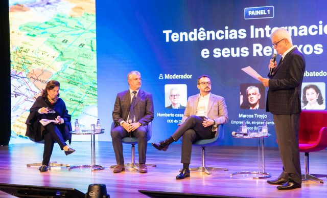 Quatro pessoas em cima de um palco, uma mulher à esquerda e três homens ao centro/ direita. A iluminação do palco é roxa e azul, todos usam vestes sociais.