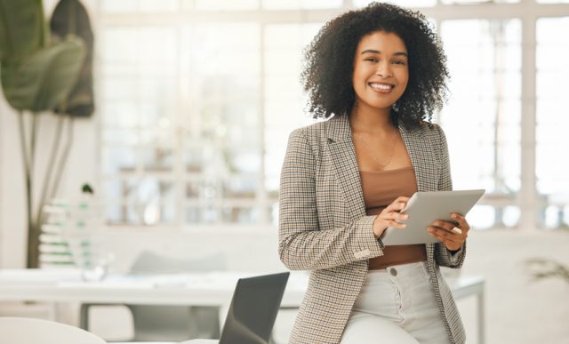 Foto de uma mulher negra sorrindo e segurando um tablet. Ela está em um escritório, usando roupas mais formais.