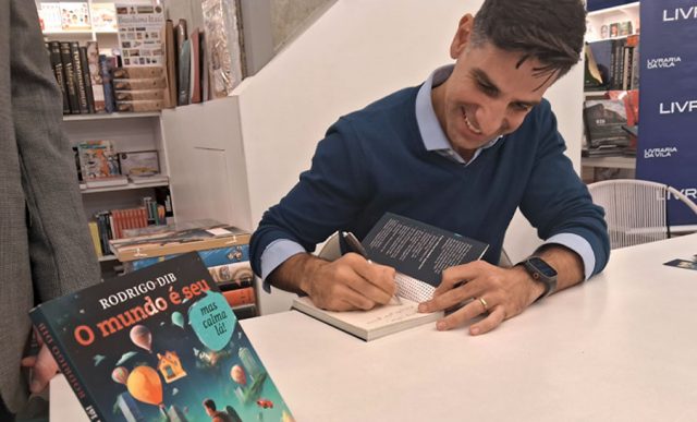 Rodrio Dib, autor do livro, escrevendo uma dedicatória na obra em cima de uma mesa branca