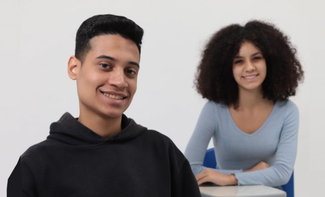 Foto com fundo cinza e dois aprendizes sentados em mesas de sala de aula olhando felizes para a câmera por saberem que agora o Programa Jovem Aprendiz Paulista contempla jovens de até 24 anos.