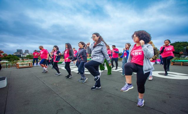 Jovens estagiários participam de uma atividade física coletiva ao ar livre. Todos usam camisetas rosa com o logo do CIEE. Eles estão em movimento, levantando uma das pernas como parte do exercício. Ao fundo, o céu está nublado e é possível ver árvores e prédios da cidade.