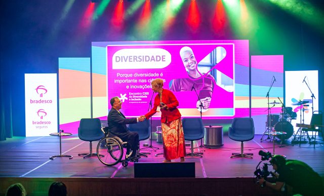 Imagem de palco com um painel ao fundo e duas pessoas ao palco, uma delas um cadeirante.