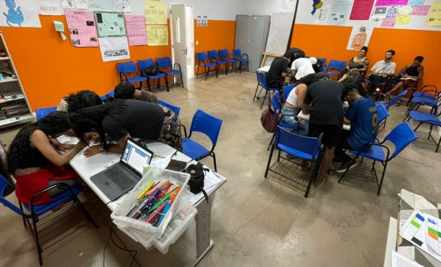 Foto de uma sala com adolescentes divididos em dois grupos realizando uma atividade e ao fundo, três pessoas observando