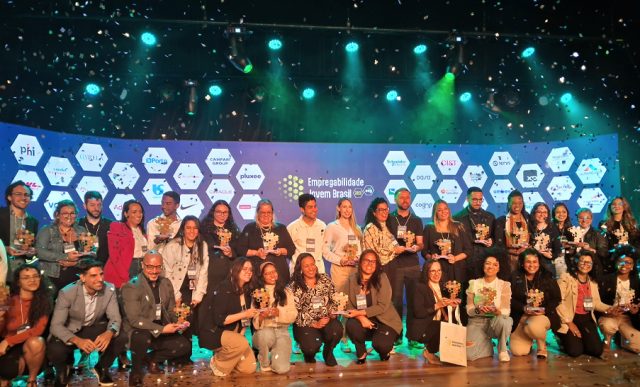 Foto de grupo registrada no palco do Teatro CIEE, em São Paulo, durante a entrega do Troféu Empregabilidade Jovem Brasil 2025. Aproximadamente 40 pessoas estão alinhadas em duas fileiras, sorrindo e segurando seus troféus, que têm o formato de peças de quebra-cabeça coloridas. No fundo, um painel azul exibe os logotipos das empresas homenageadas. Confetes prateados estão caindo, registrando o clima de celebração. A iluminação do palco é azul esverdeada, dando destaque ao cenário.