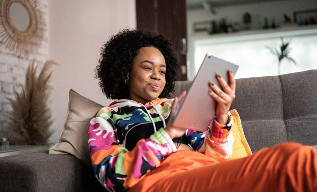 Moça negra sentada ao sofá navegando na internet com um tablet