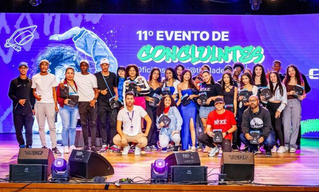 Jovens em um palco, posando para foto, atrás há um telão com o nome do evento e fundo roxo