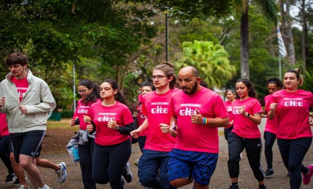 Fotografia de um grupo de pessoas correndo em um parque arborizado. Todos vestem camisetas rosa com a inscrição “Treino Exclusivo CIEE” na frente. Na linha de frente, três homens e três mulheres correm concentrados, com expressões de foco e determinação. Ao fundo, outras pessoas participam da corrida em meio às árvores.
