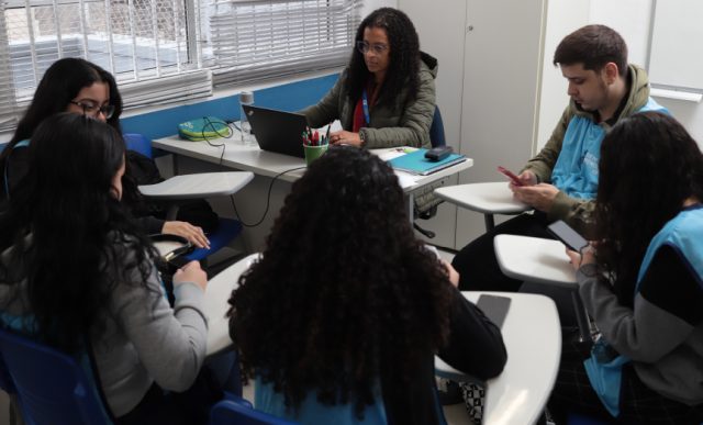 Grupo de jovens sentados em uma roda de estudo com celular e notebook em mãos.