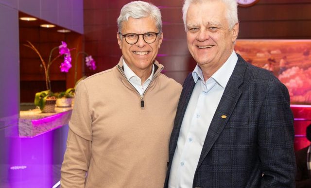 Dois homens estão em pé e sorrindo para a câmera. À esquerda está Robert John van Dijk, de cabelos grisalhos e óculos, vestindo suéter bege. À direita está Humberto Casagrande, de cabelos brancos, vestindo blazer azul escuro e camisa social azul clara. Ao fundo, parede de madeira com uma pintura e um relógio redondo.