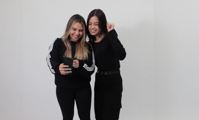 Foto de duas estudantes brancas, vestidas com roupas na cor preta, olhando para o celular e comemorando a busca por uma vaga de estágio no CIEE.
