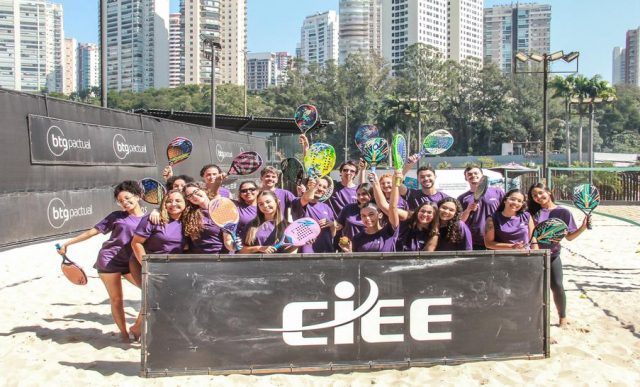 Grupo de jovens estagiários em uma quadra de areia, todos usando camisetas roxas. Eles posam sorridentes atrás de uma placa preta com o logotipo do CIEE em branco, segurando raquetes de beach tênis.